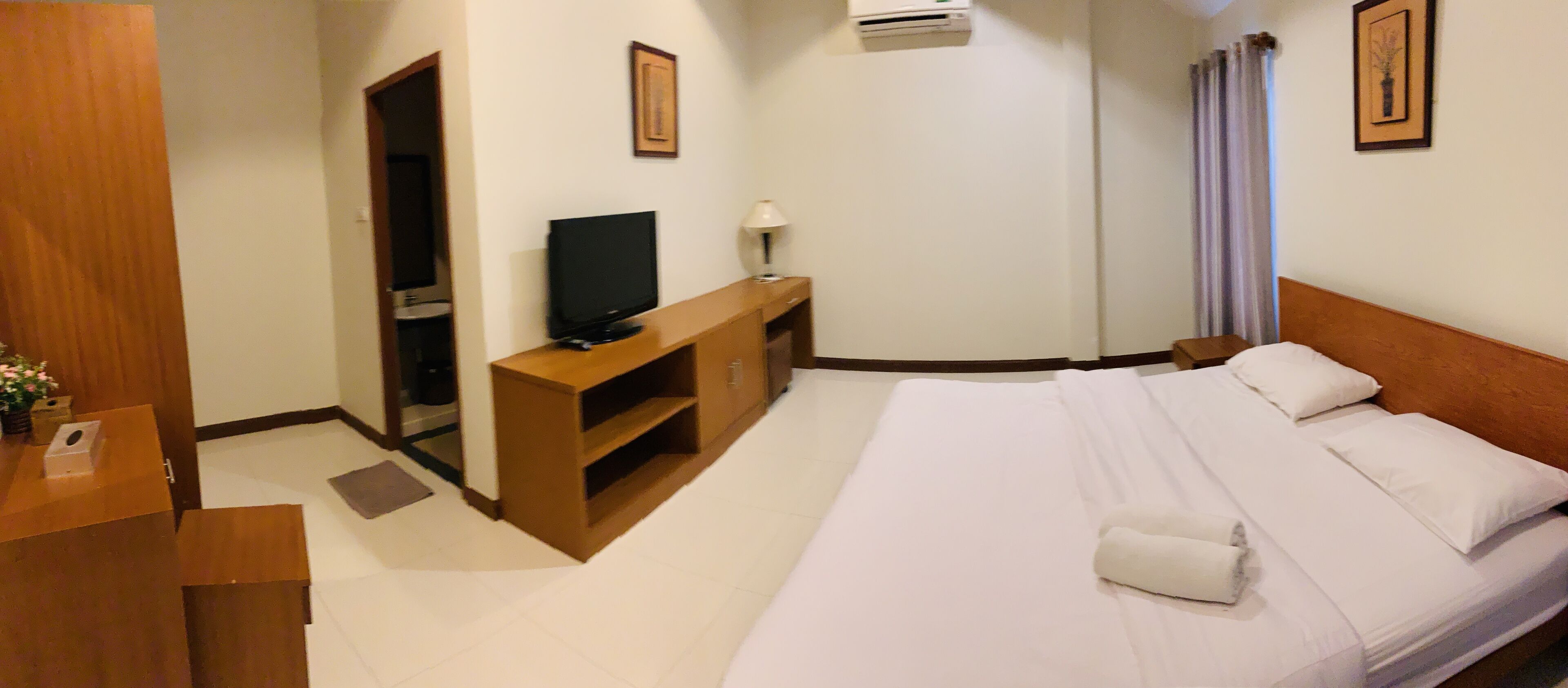 Una habitación de hotel con una cama, un televisor sobre un soporte de madera, un escritorio y un baño visible a través de una puerta abierta.