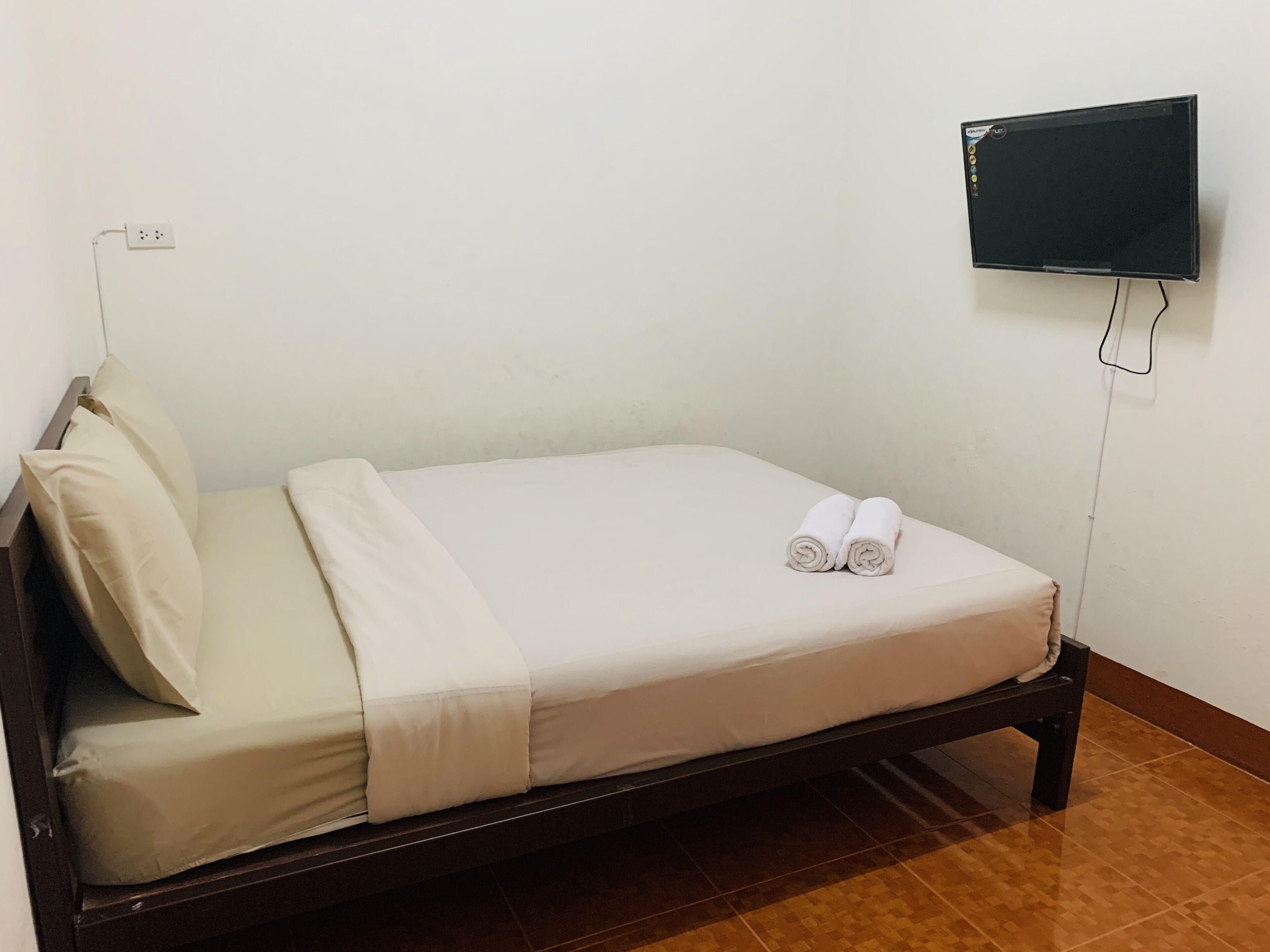 Een eenpersoonsbed met een witte matras, een beige deken en twee opgerolde handdoeken. Een flatscreen televisie is rechts op de muur bevestigd.