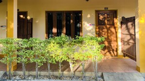 A building entrance with a yellow facade, two wooden doors, a security camera, and a small garden area with young trees. - Srisupawadee Resort (Prachuap Khiri Khan)