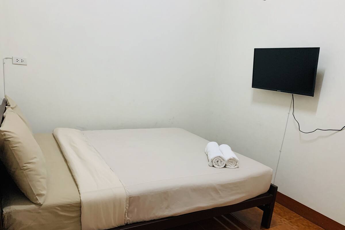 Ein Einzelbett mit weißer Matratze und beigen Kissen, ein an der Wand befestigter Fernseher und eine Steckdose an der Wand.