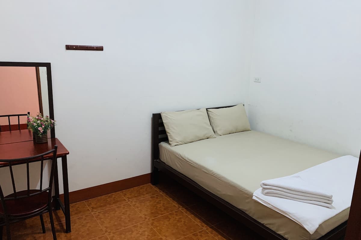 Ein kleines Zimmer mit einem Einzelbett, einem Holztisch und einem Stuhl, einer kleinen Topfpflanze und einer weißen Wand.