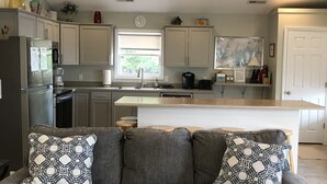 Een moderne keuken met een grijze bank en gekleurde kussens, een inbouw kookeiland, en een raam met uitzicht op groen.