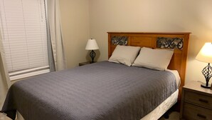 Een opgemaakt bed met een grijze deken, een houten hoofdbord met decoratieve metalen details, twee bedlampen en een klein houten nachtkastje.
