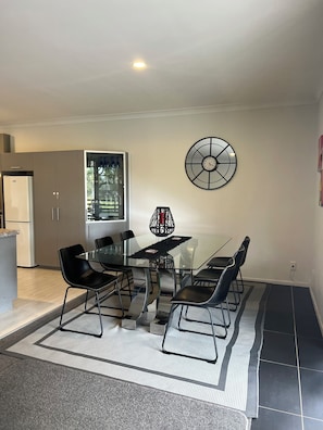 A modern dining area with a glass table and black chairs, a black clock on the wall, and a kitchen in the background. - A Cut Above - gateway to the Bay of Islands (Kerikeri)