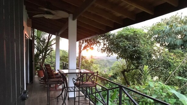 A covered outdoor seating area with a table and chairs, overlooking a lush garden and a distant view of the landscape. - Villas Lambeau #2 - Ocean View - Fiber Optic / 1BR 1 BA (Uvita)