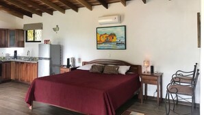 A bedroom with a bed, a nightstand, a chair, a painting, and a kitchen area with cabinets and a refrigerator. - Villas Lambeau #2 - Ocean View - Fiber Optic / 1BR 1 BA (Uvita)