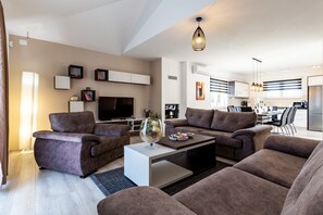 En moderne stue med to sofaer, et sofabord og et fjernsyn. I baggrunden er der et køkkenområde med et spisebord og stole.