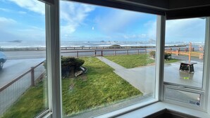 A view from a room with large windows overlooking a grassy area, a road, and a coastal scene with waves and a bridge. - Oceanfront Retreat. Sunset Views. Whale Watching. Redwood Groves, 2 King Beds (Crescent City)