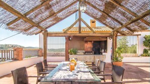 An outdoor patio with a glass table set for dining, surrounded by wicker chairs, under a thatched roof structure. - Lovely home in Iznájar (Iznájar)