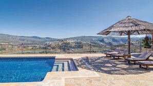 A pool with a stone border, lounge chairs, a thatched umbrella, and a view of a hilly landscape. - Lovely home in Iznájar (Iznájar)