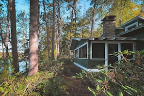 A cabin with a stone chimney, surrounded by tall trees and a forested area. - Premier Cabin 1 with Lake View (Paul Smiths)