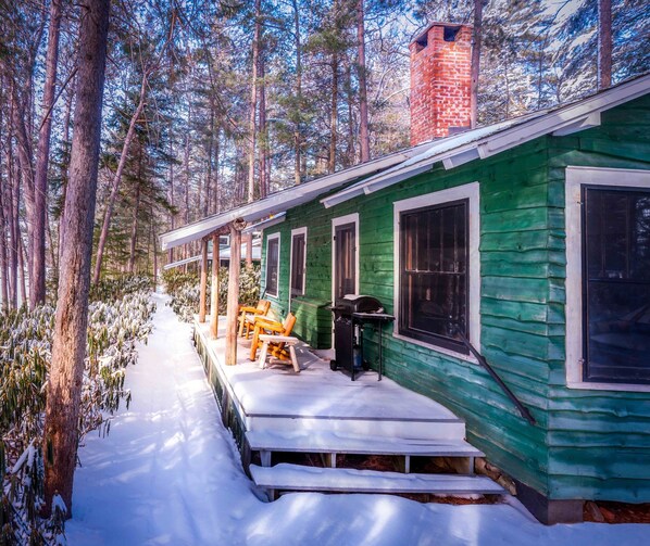 A green wooden cabin with a porch, steps leading up to the entrance, and a chimney. - Premier Cabin 1 with Lake View (Paul Smiths)