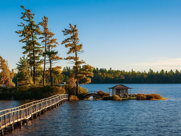 A wooden pier extending into a lake, surrounded by tall trees and a small gazebo on an island.