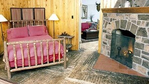 A cozy room with a bed, a stone fireplace, and wooden walls. - Premier Cabin 1 with Lake View (Paul Smiths)