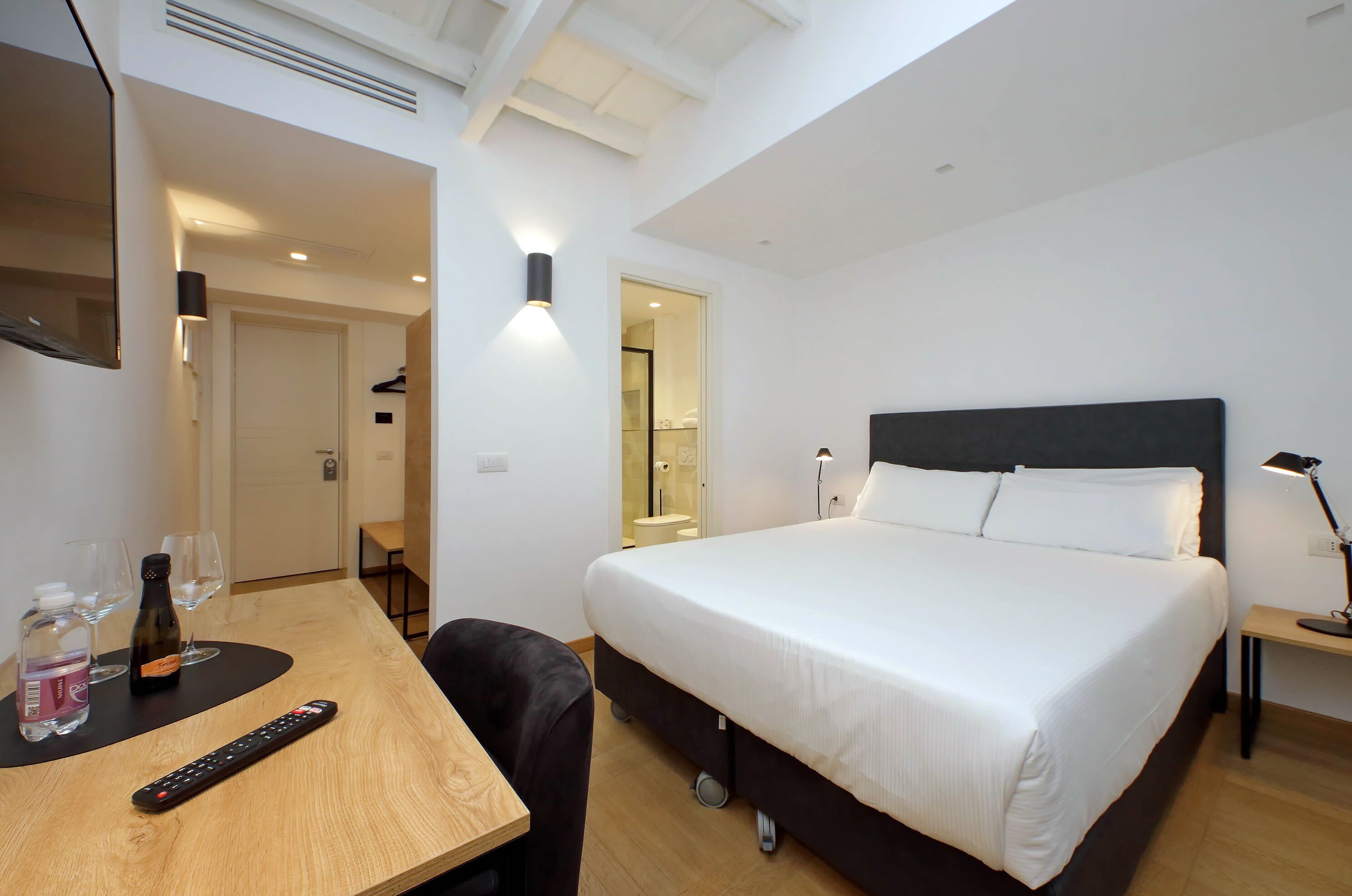 A hotel room with a bed, a desk with a remote control, a bottle, and glasses.