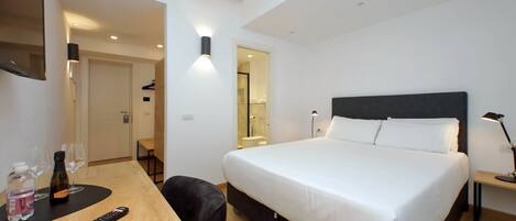 A hotel room with a bed, a desk with a remote control, a bottle, and glasses.