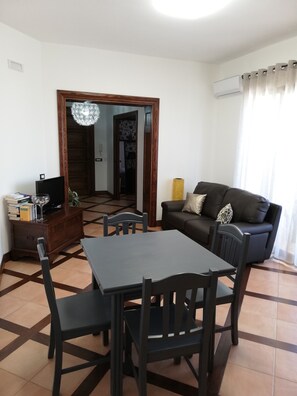 A living room with a dining table, chairs, a sofa, a TV on a wooden cabinet, and a patterned wallpaper door.
