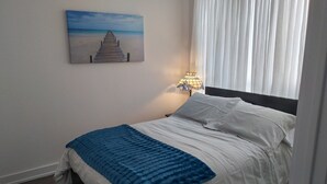 A neatly made bed with white and blue bedding, a painting of a beach scene on the wall, and a lamp on the nightstand.