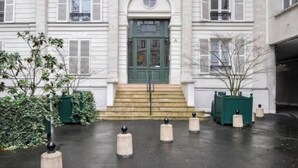 A building with a central entrance, a staircase leading up to it, and a paved area with bollards.