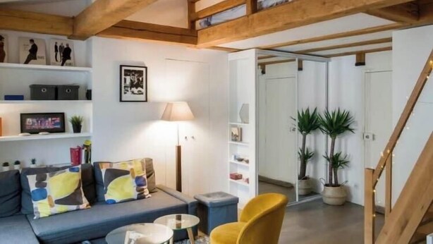 A modern living room with a wooden ceiling, a sofa with colorful cushions, a small table, and a yellow chair. There are shelves with various items and a lamp.