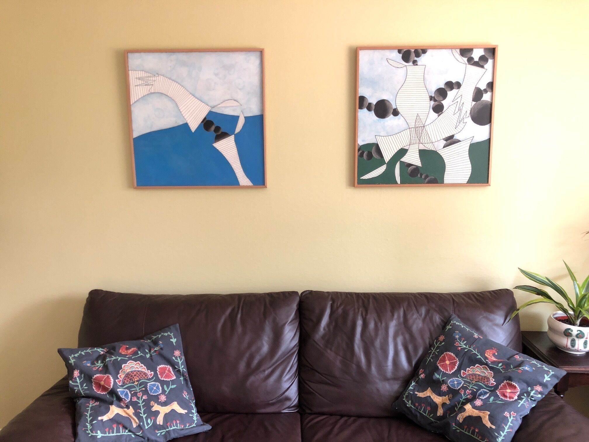 Un divano in pelle marrone con due cuscini decorativi, un quadro astratto incorniciato al muro e una pianta in vaso su un tavolino.
