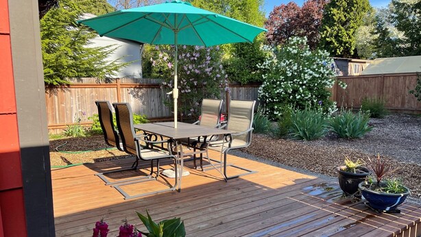 Una terraza de madera con una mesa, sillas, un parasol verde y plantas en macetas.