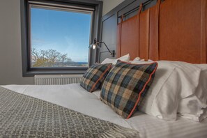 Ein ordentlich bezogenes Bett mit Karo-Kissen, ein Fenster mit Ausblick und eine Wandleuchte.