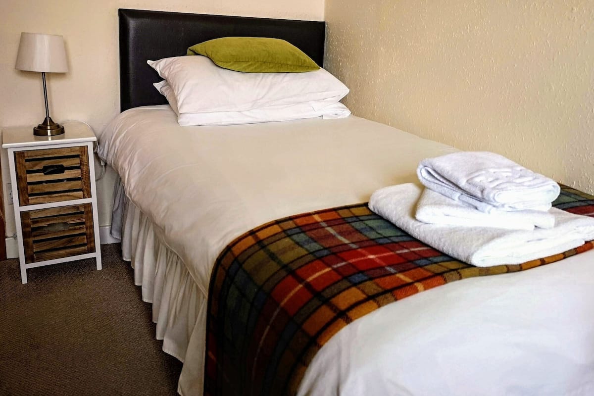 A neatly made bed with white bedding and a plaid blanket, a small bedside table with a lamp, and a wooden storage unit.