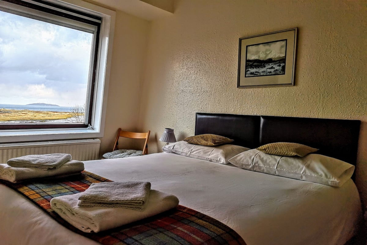 A neatly made bed with pillows and folded towels, a framed picture on the wall, and a view of the outdoors through a window.