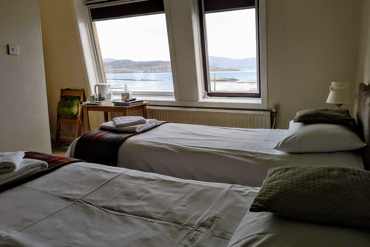 A hotel room with two beds, a view of the sea, a table with a lamp, and a chair.