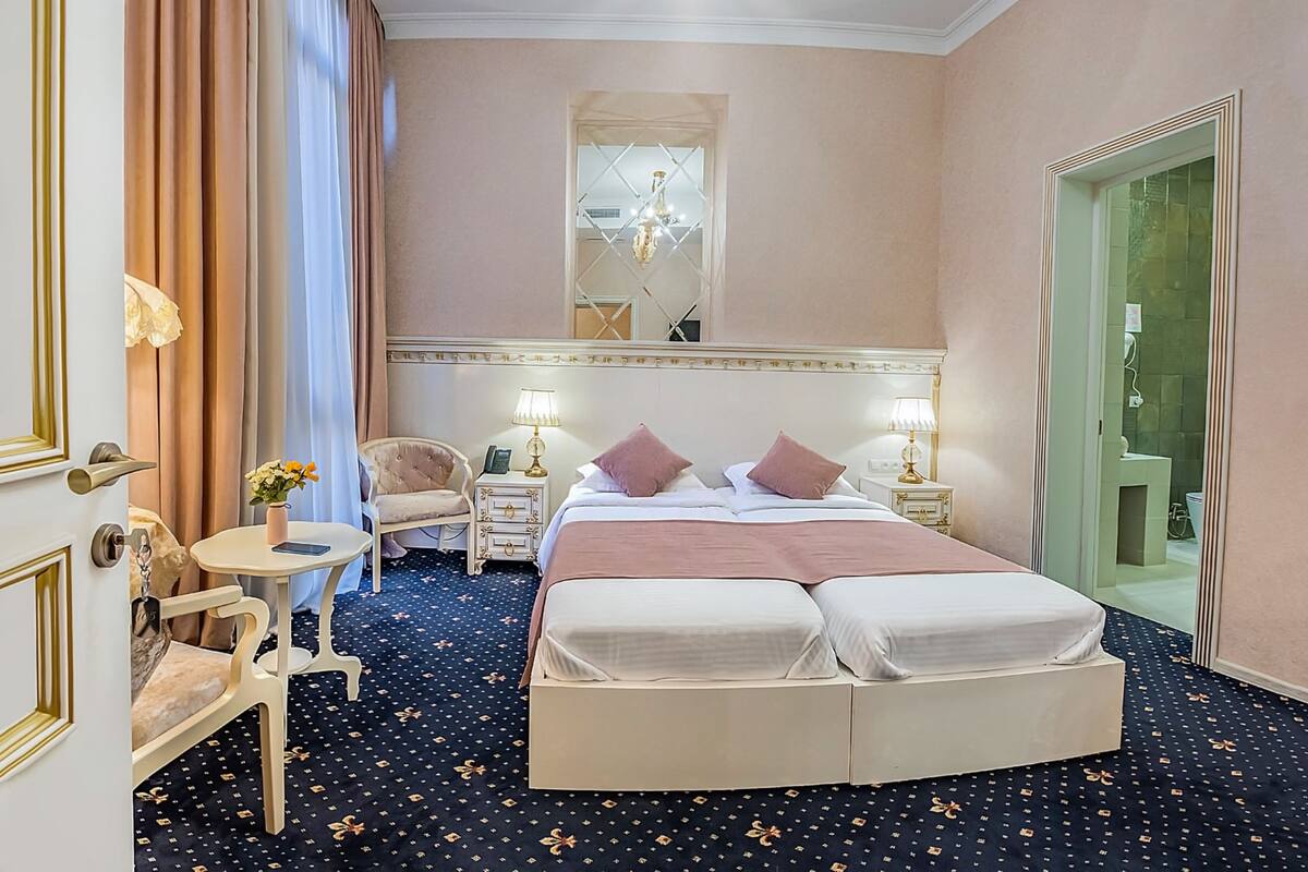 A hotel room with a large bed, two bedside tables with lamps, a chair, a small table with a vase, and a mirror above the bed.