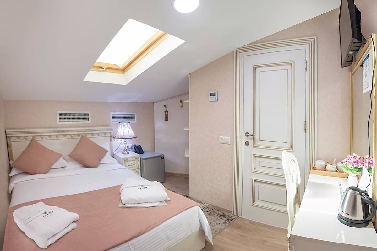 A hotel room with a bed, bedside table, lamp, and a desk with a kettle.