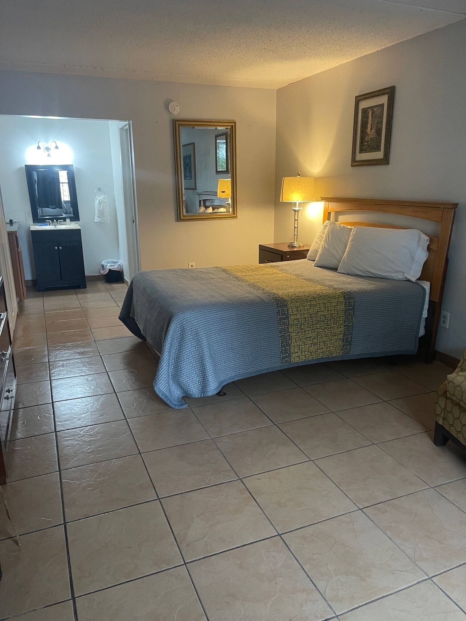 A hotel room with a bed, a nightstand, a lamp, a mirror, a framed picture, and a bathroom visible in the background.