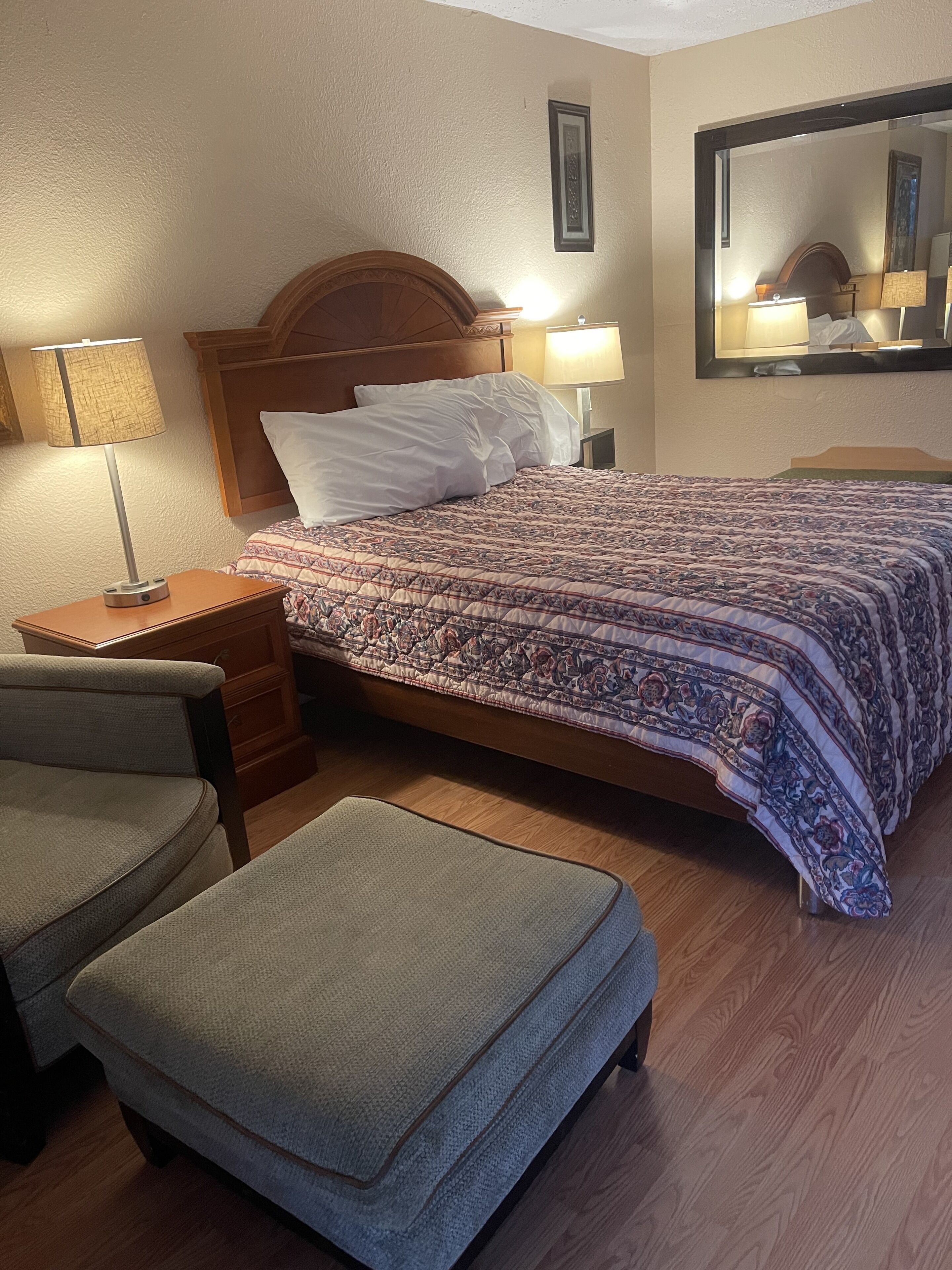 A hotel room with a bed, two bedside lamps, a chair, an ottoman, and a mirror on the wall.