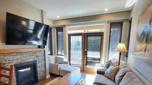Ein modernes Wohnzimmer mit Flachbildfernseher an der Wand, Kamin, einer Couch, einem Couchtisch und einem Essbereich mit Holztisch und Stühlen.