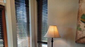 Ein Wohnzimmer mit einem beigefarbenen Sofa, einem Sessel und einer Lampe. Große Fenster mit Jalousien und ein Gemälde an der Wand.
