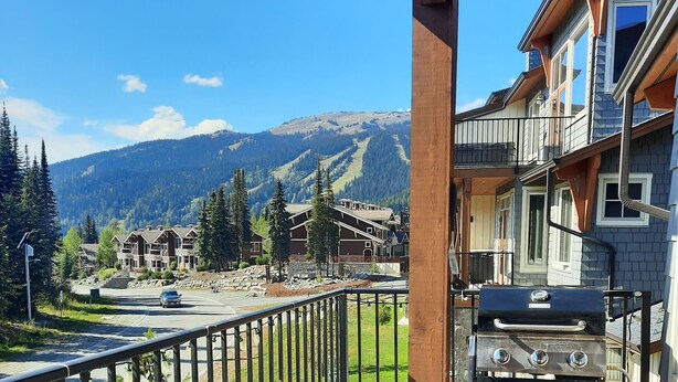 Ein Balkon mit Korbmöbeln und einem Grill, mit Blick auf eine gebirgige Landschaft und ein Wohngebiet.