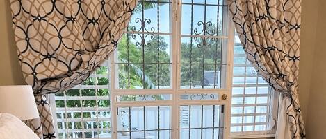 A bedroom with a bed, curtains, a window with a view of greenery, and a door with a metal gate.