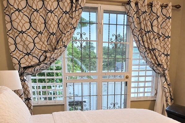 A bedroom with a bed, curtains, a window with a view of greenery, and a door with a metal gate.