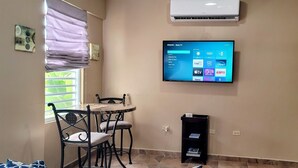 A small room with a TV mounted on the wall displaying various streaming services. There is a window with blinds, a table with chairs, and a small cabinet.