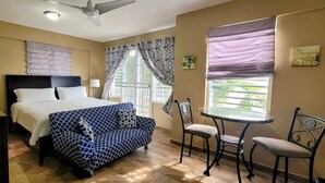A bedroom with a bed, a desk, two chairs, a ceiling fan, and a window with curtains.
