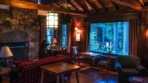 Un intérieur chaleureux de chalet avec une cheminée en pierre, des poutres en bois, une grande fenêtre donnant sur la forêt et un canapé à carreaux rouges.