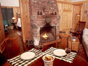 A cozy room with a brick fireplace, wooden furniture, and a dining table set for two. - Signature Cabin 2 with Lake View (Paul Smiths)