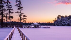 Un sentier enneigé mène à une petite construction en bois, surmontée d’un toit, et qui est entourée de grands arbres et d’un lac gelé.