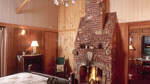 Une pièce chaleureuse avec une cheminée en briques, des murs en bois, un lit recouvert d’une courtepointe à motifs et une petite table avec une lampe.