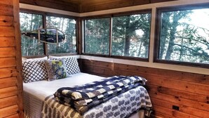 Una habitación cálida tipo cabaña con cama, ventanas amplias y vista a árboles.