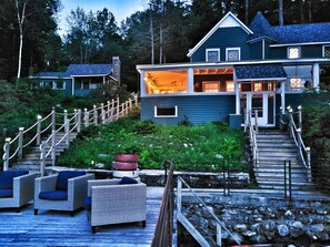 Une maison à deux étages dotée d’une terrasse en bois, d’un espace salon extérieur et d’un escalier en pierre menant à l’entrée.