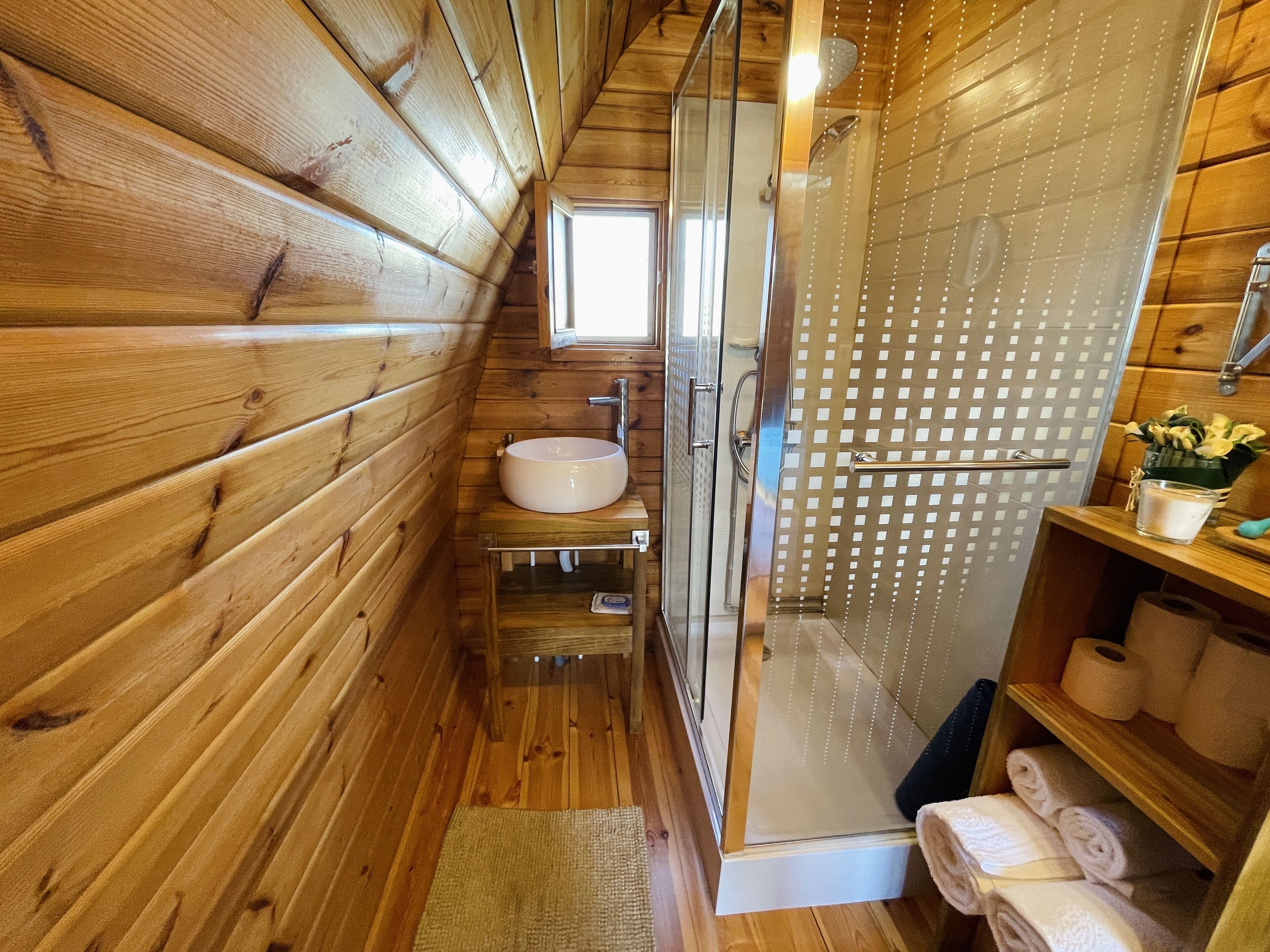A wooden cabin bathroom with a glass-enclosed shower, a sink, and a shelf with towels.