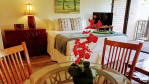 A hotel room with a bed, a TV, a wooden dresser, a lamp, and a table with a vase of flowers. - Oceanfront, King Bed, AC, Wi-Fi, Hot Tub, Pool (Lahaina)