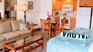 A compact living space with a sofa, a glass coffee table, a dining table with chairs, a kitchenette with a microwave and refrigerator, and a ceiling fan. - Oceanfront, King Bed, AC, Wi-Fi, Hot Tub, Pool (Lahaina)